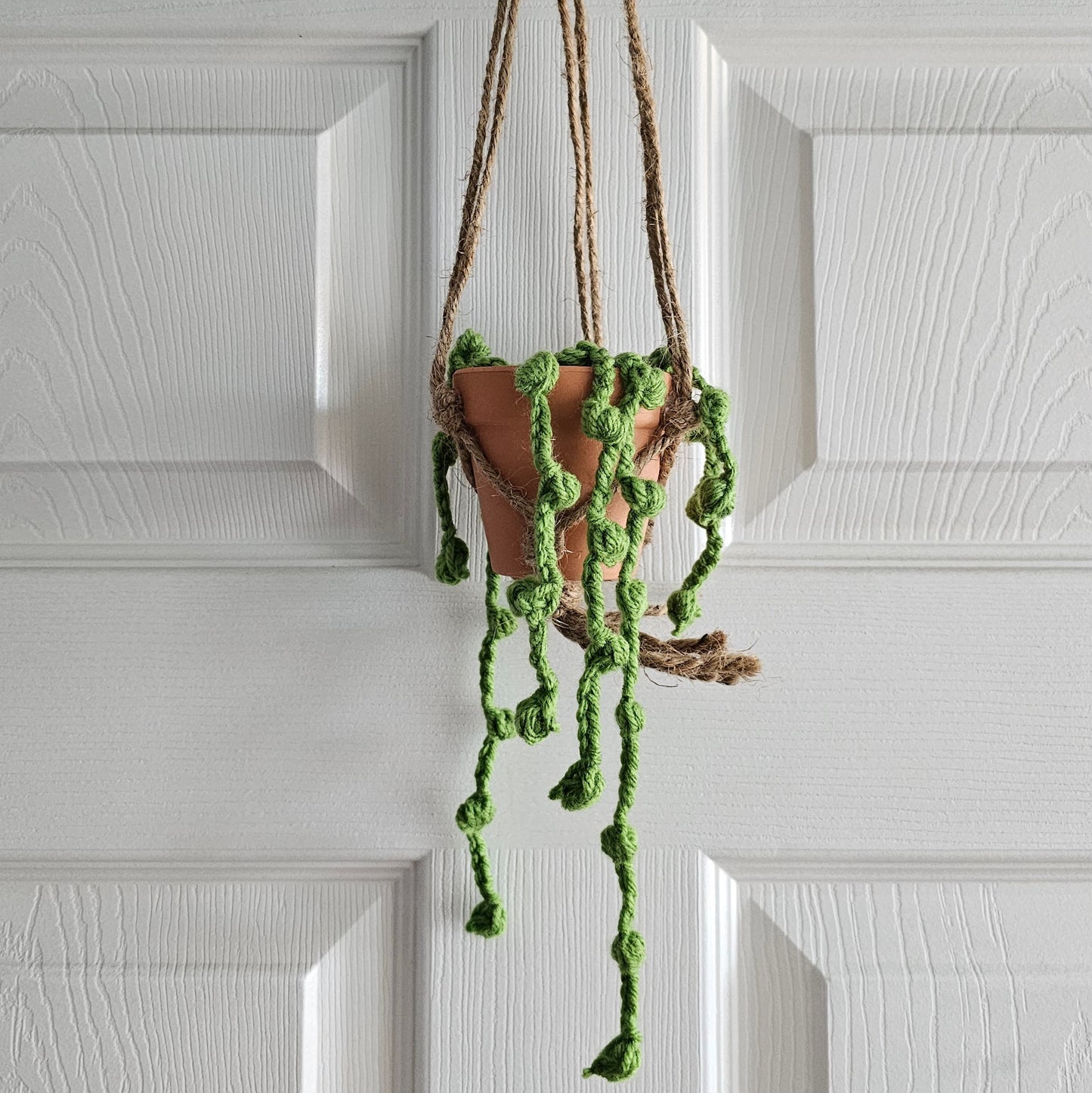 String of pearls plant crochet from light green yarn, hangs from a suspended terracotta pot. This pot sits in a jute rope macrame in front of a white background.