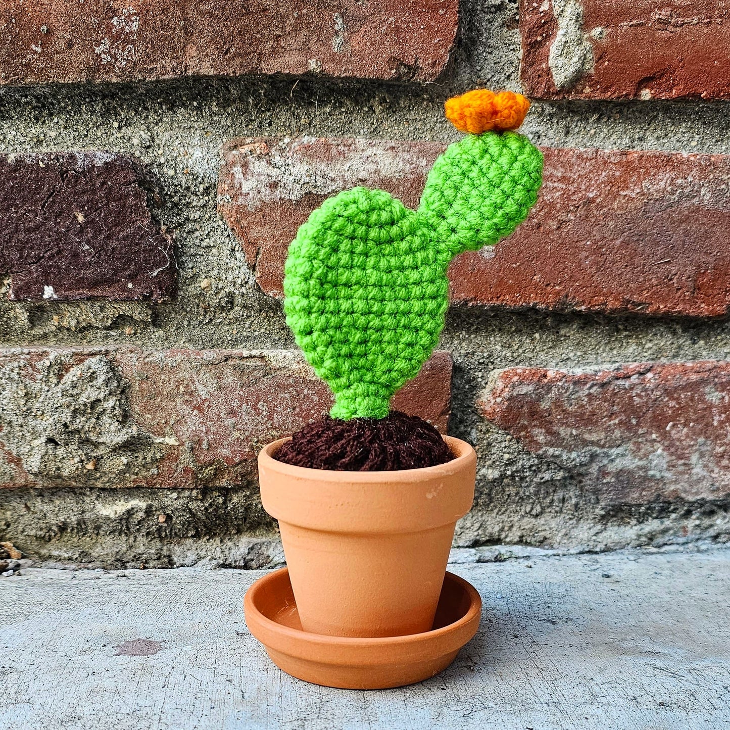 Crochet bright green prickly pear cactus with an orange flower growing out of the top. The cactus is growing out of crochet dirt made out of brown yarn. The cactus sits in a small terracotta clay pot and saucer.