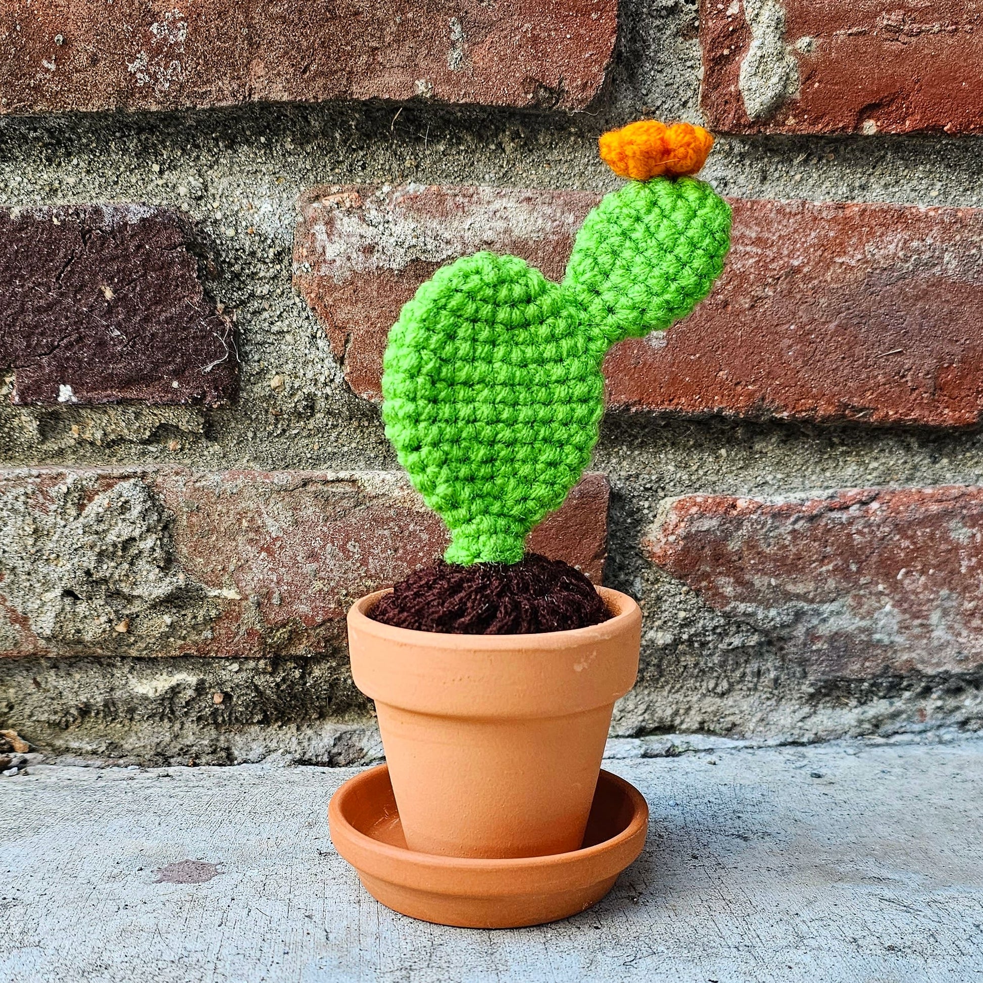 Crochet bright green prickly pear cactus with an orange flower growing out of the top. The cactus is growing out of crochet dirt made out of brown yarn. The cactus sits in a small terracotta clay pot and saucer.
