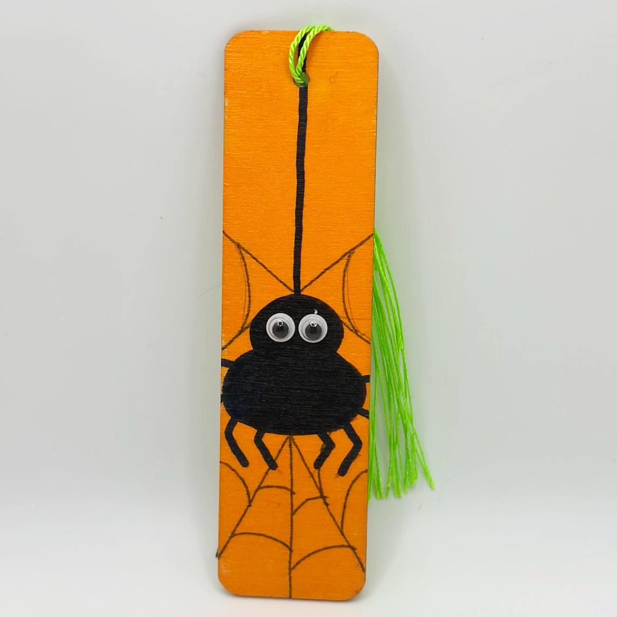 Small, orange, wooden bookmark decorated with a drawn on black spider with googly eyes, sitting on a web. The bookmark has a bright green tassel attached at the top through a small hole.