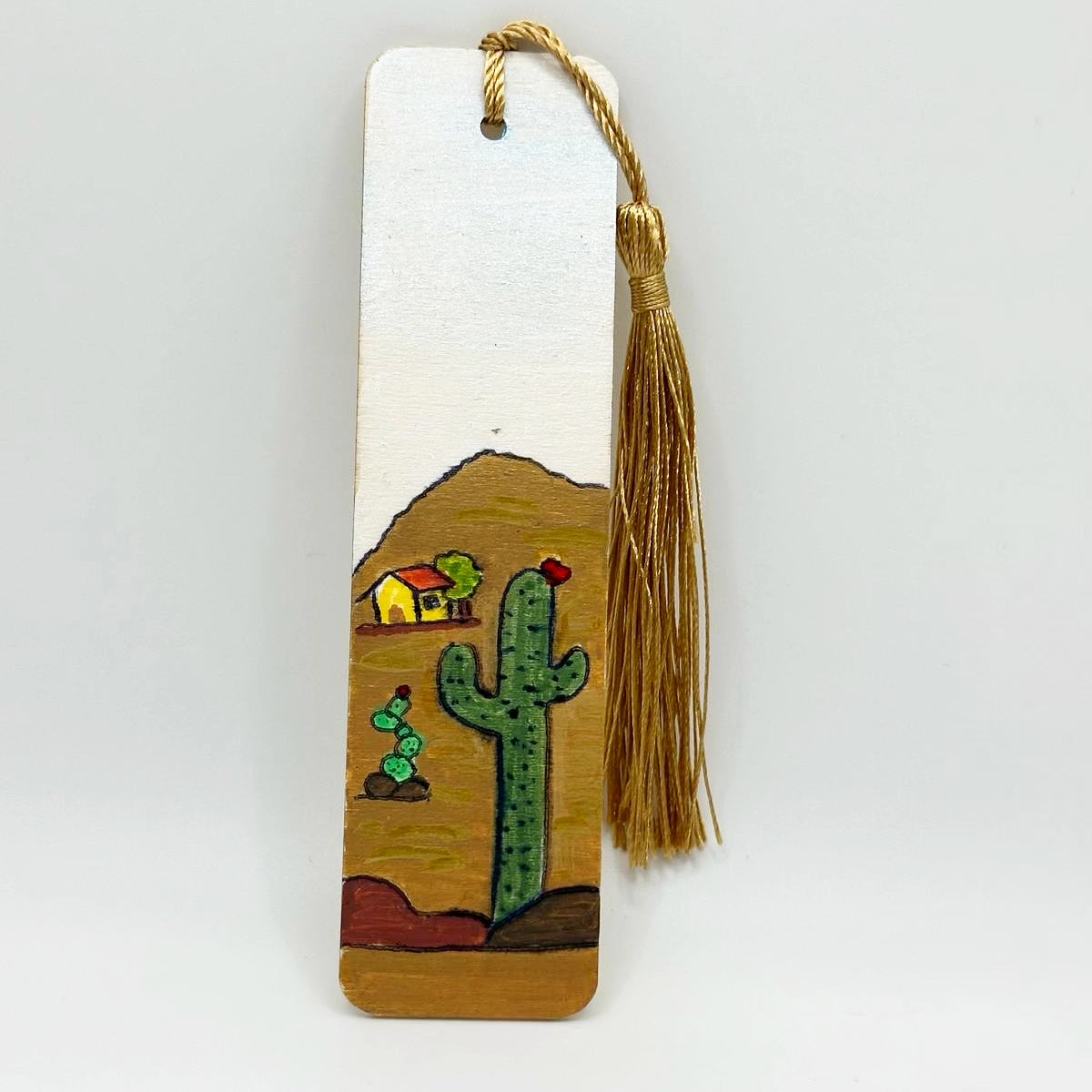 Small wooden bookmark decorated with a picture of a landscape of a mountain. On the mountain there is a small yellow house with an orange roof and a little tree beside it. Growing out of small mounds of dirt are two separate cacti which are covered in thorns and are topped with a small cute red flower. There is a golden tassel attached to the bookmark through a hole at the top.