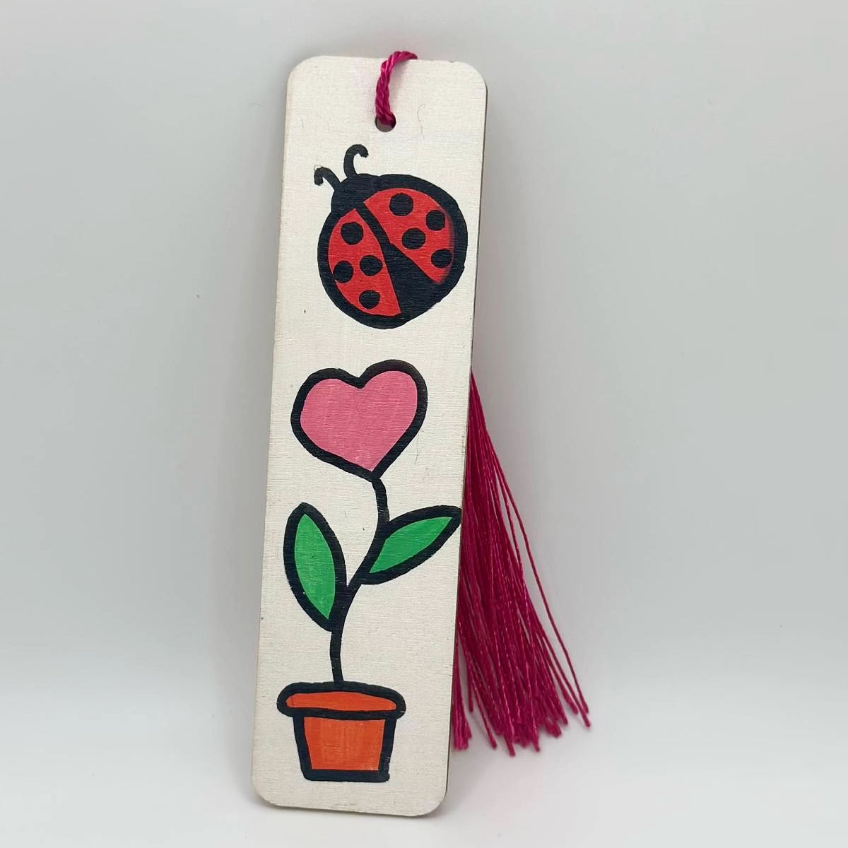 Small, white, wooden bookmark decorated with a pink heart shaped flower growing out of an orange flower pot. There is a red and black ladybug with small black dots on its wings floating near the flower. The The bookmark has a pink tassel attached at the top through a small hole.