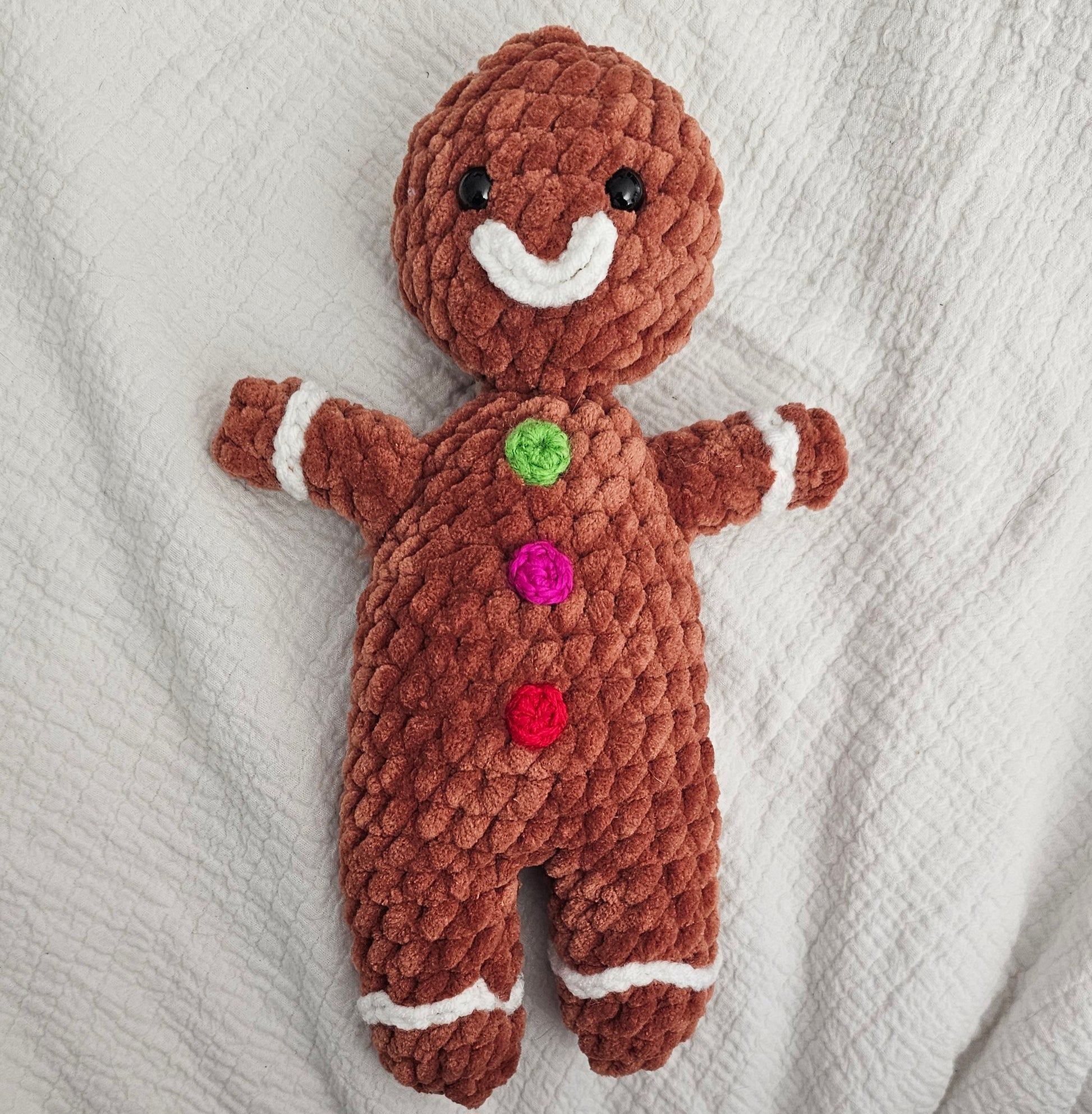 Gingerbread man made from brown plush yarn. It has white frosting lips and cuffs at the wrists and ankles made from smaller white yarn. This gingerbread man has three colorful buttons (fuchsia, red and bright green).