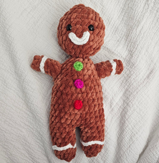 Gingerbread man made from brown plush yarn. It has white frosting lips and cuffs at the wrists and ankles made from smaller white yarn. This gingerbread man has three colorful buttons (fuchsia, red and bright green).