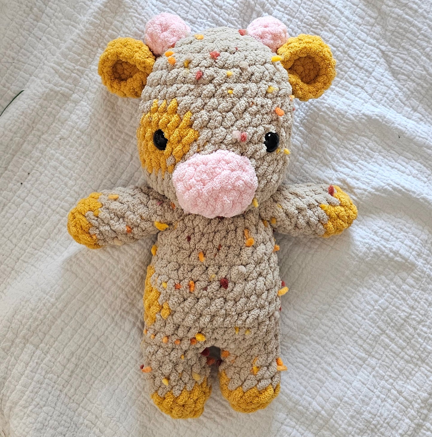 Cow plushie made from light brown yarn with orange and yellow sprinkles. The cow has a marigold yellow spot on its left eye and side of the body. The ears, hooves and tail are marigold yellow color as well. the cow has black eyes and pink snout and horns.