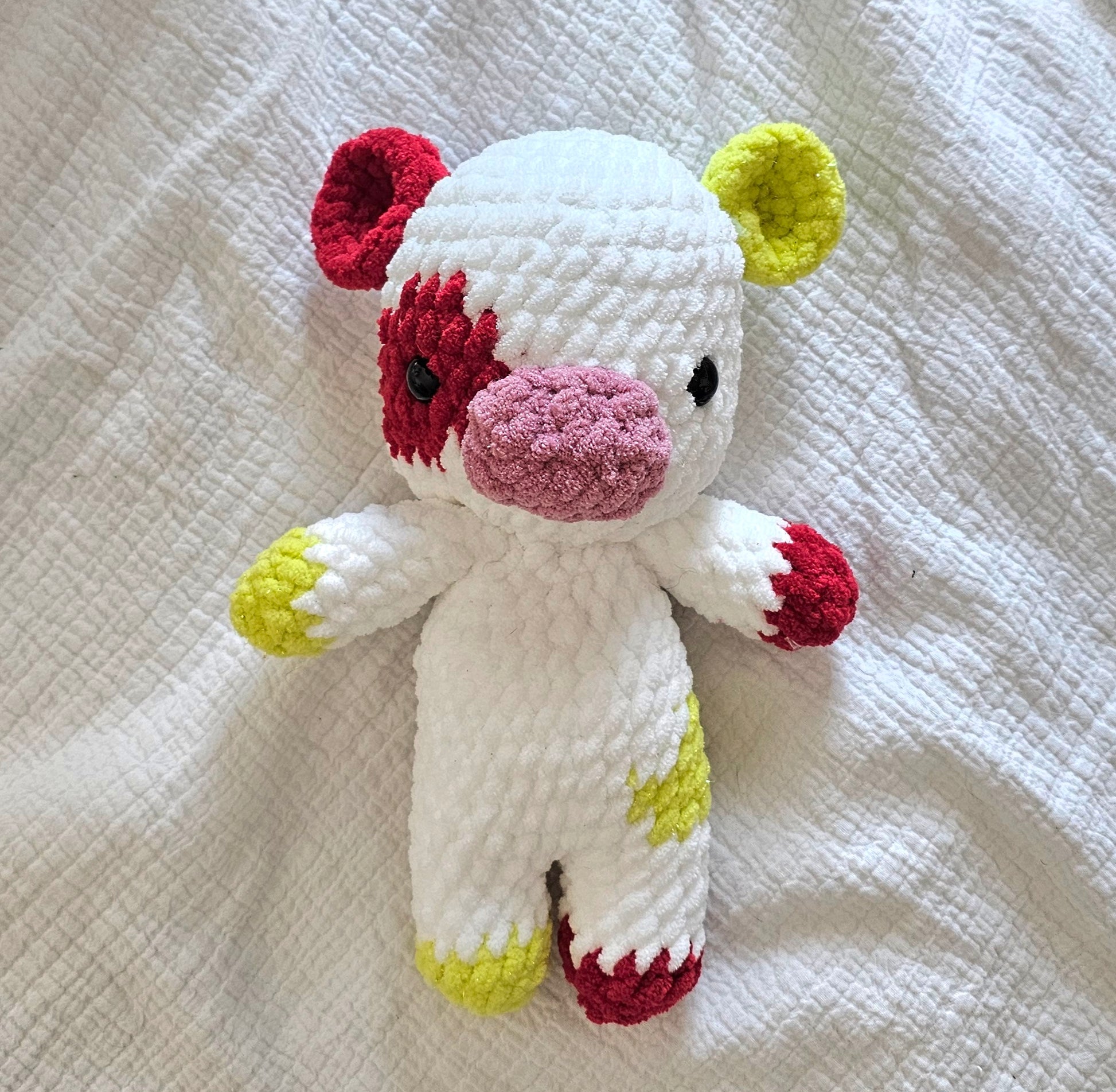 Cow plushie is made from soft white plush yarn. The ears, hooves, and tail alternate between bright sparkly green and red. The cow has a red spot on its left eye and a green spot on the right leg. It has a pink snout.