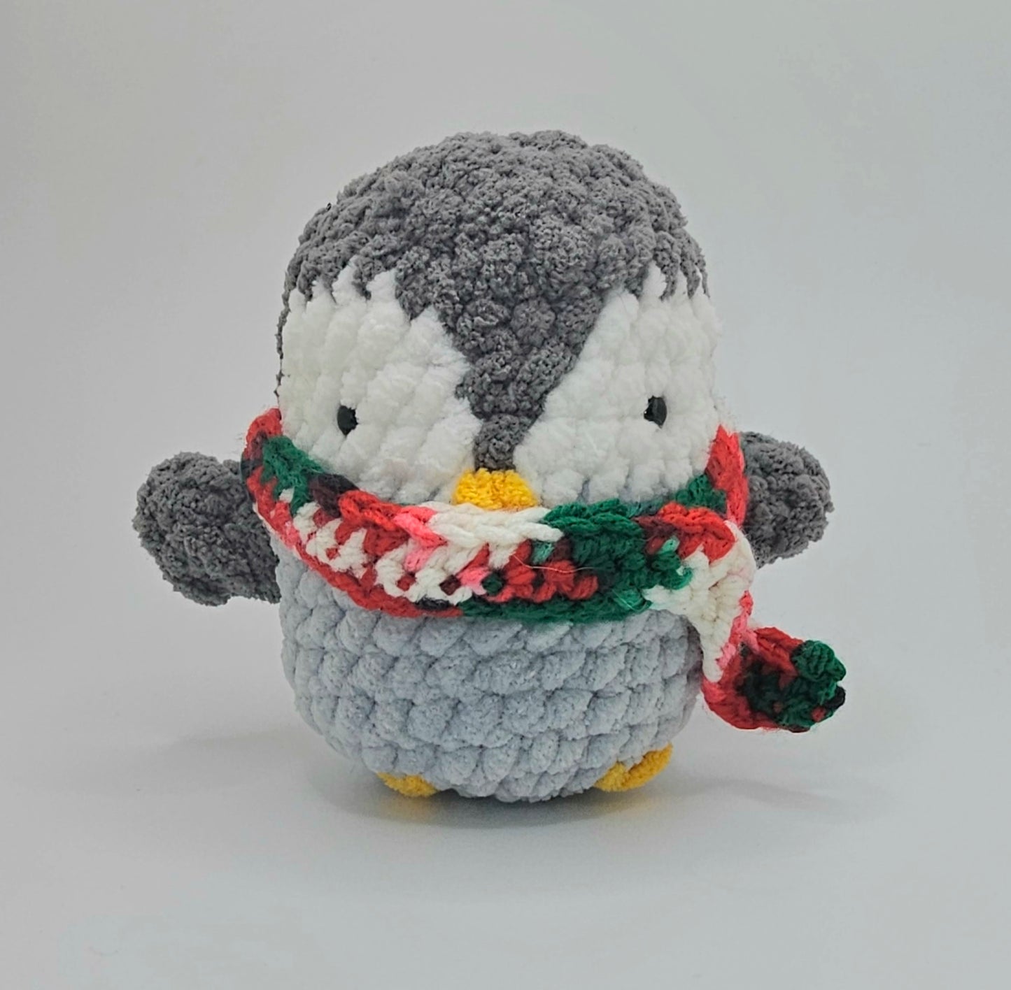 Small plushie penguin made from three different colors of yarn. The bottom half of the penguin is a light gray. The area around the eyes is white yarn, and the rest is a darker gray color including the wings. The beak and feet are made from bright yellow yarn. The penguin is wearing a green, white and red scarf.