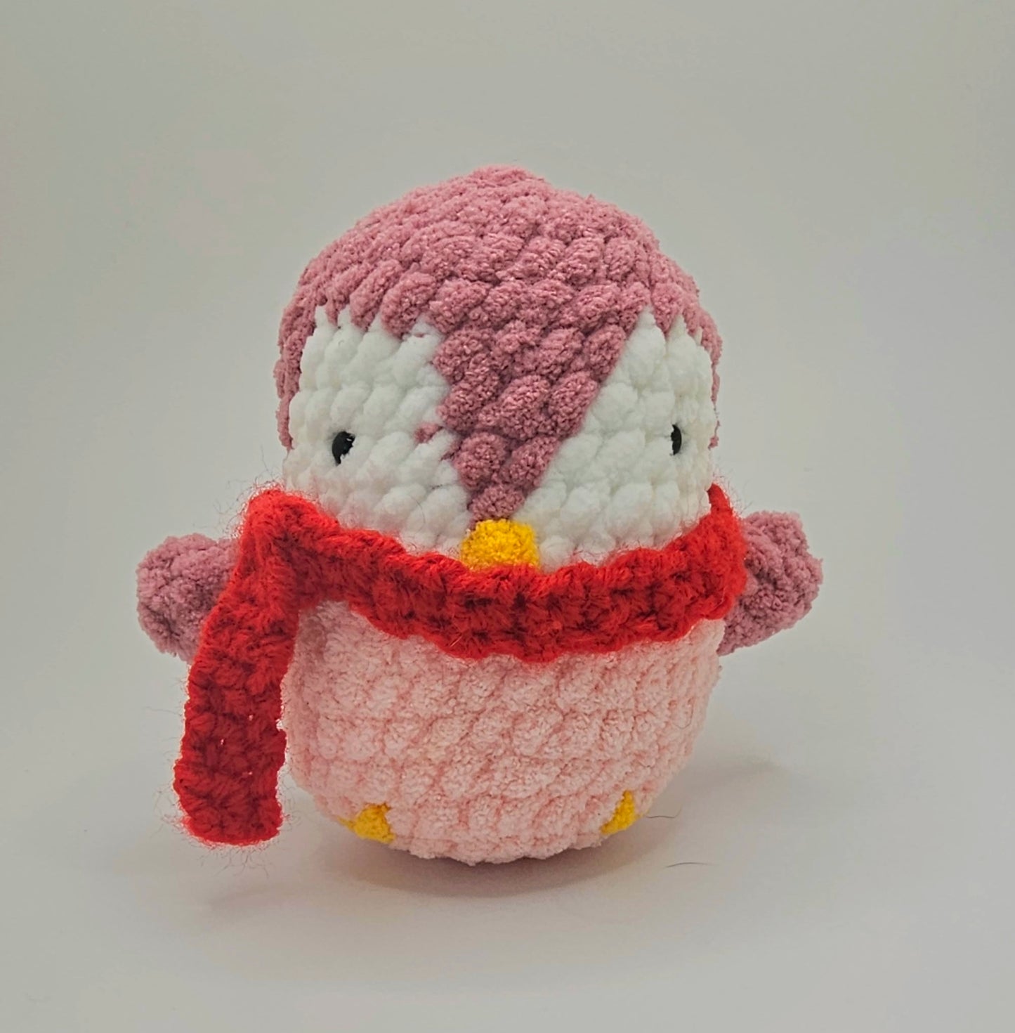 Small plushie penguin made from three different colors of yarn. The bottom half of the penguin is a baby pink, the area around the eyes is white and the rest of the penguin is a mauve color including the wings. The beak and feet are made from a bright yellow yarn. The penguin wears a bright red crochet scarf.