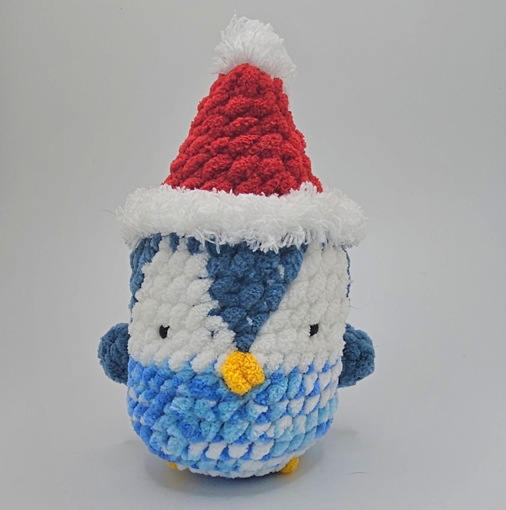 Small plushie penguin made from three different colors of yarn. The bottom half of the penguin is a color blend of dark blue, light blue and white, the area surrounding the eyes is white and the top of the penguin and the wings are a darker blue color. The beak and feet are made out of bright yellow yarn. The penguin is wearing a Santa hat made out of red yarn and white fuzzy yarn lining the top and bottom of the hat.