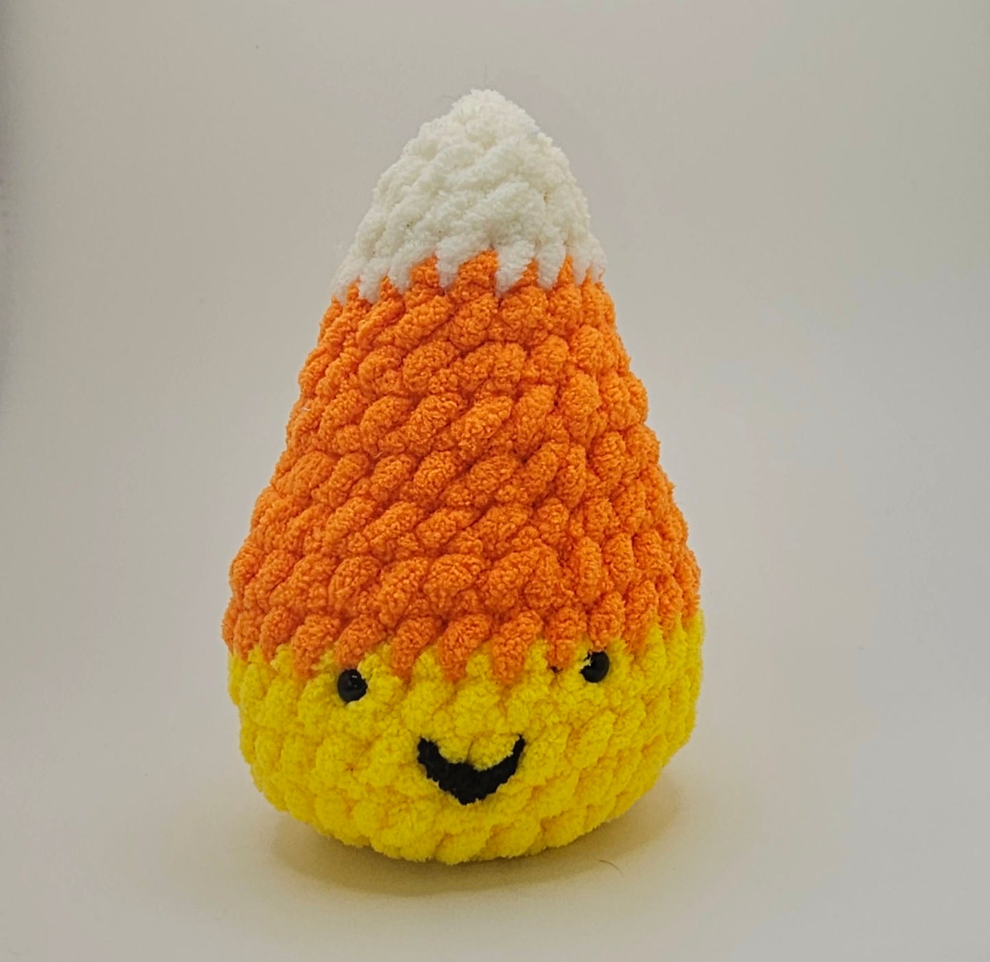 Candy corn plushie made out of three different colored yarn. Yellow at the base, orange in the middle, and white on the top. The candy corn plushie has two small black eyes and a small smiley mouth made out of black yarn