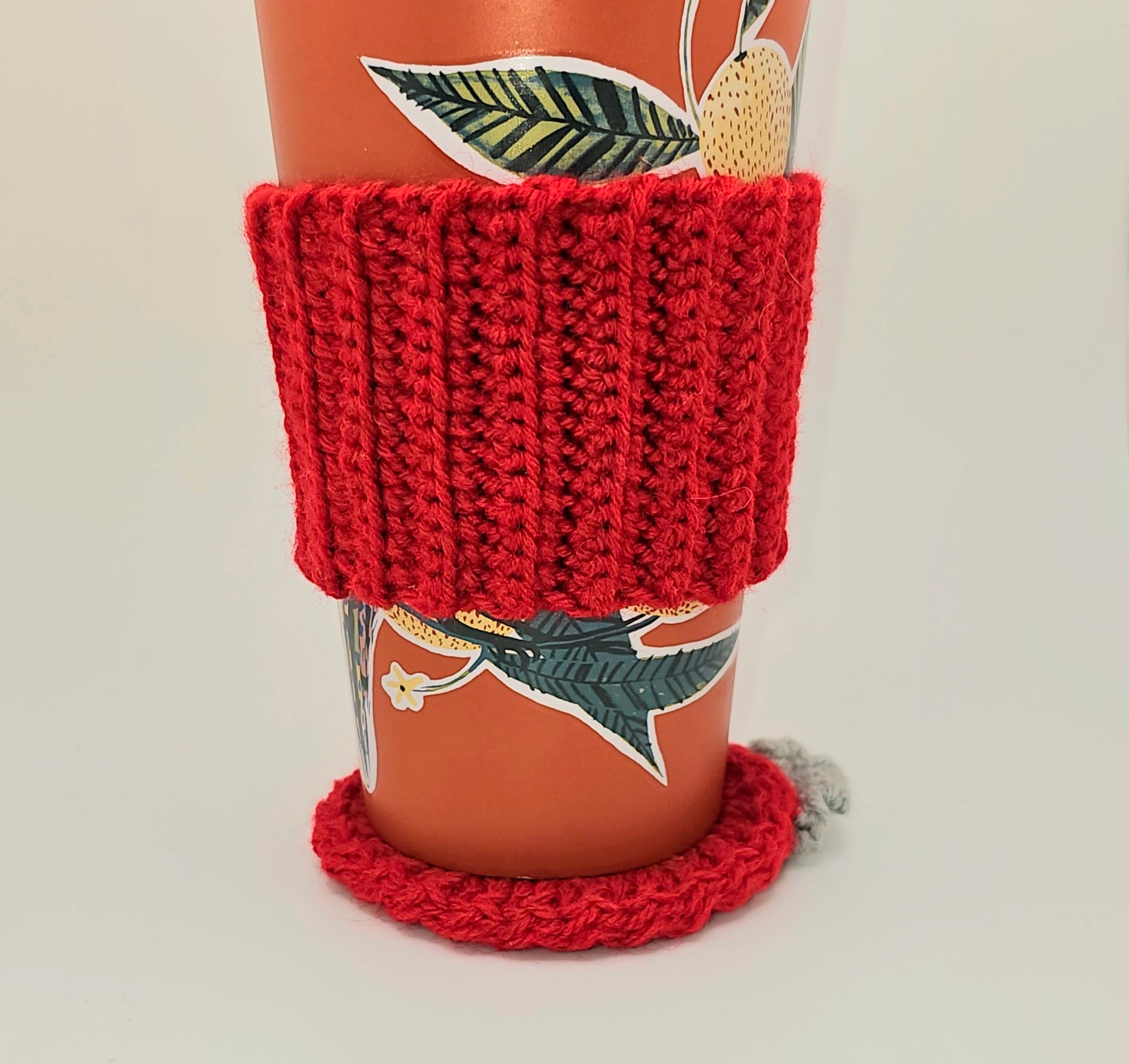 Red coffee cozy hugging a handle-less orange coffee cup. The cup also sits on top of a crochet red coaster