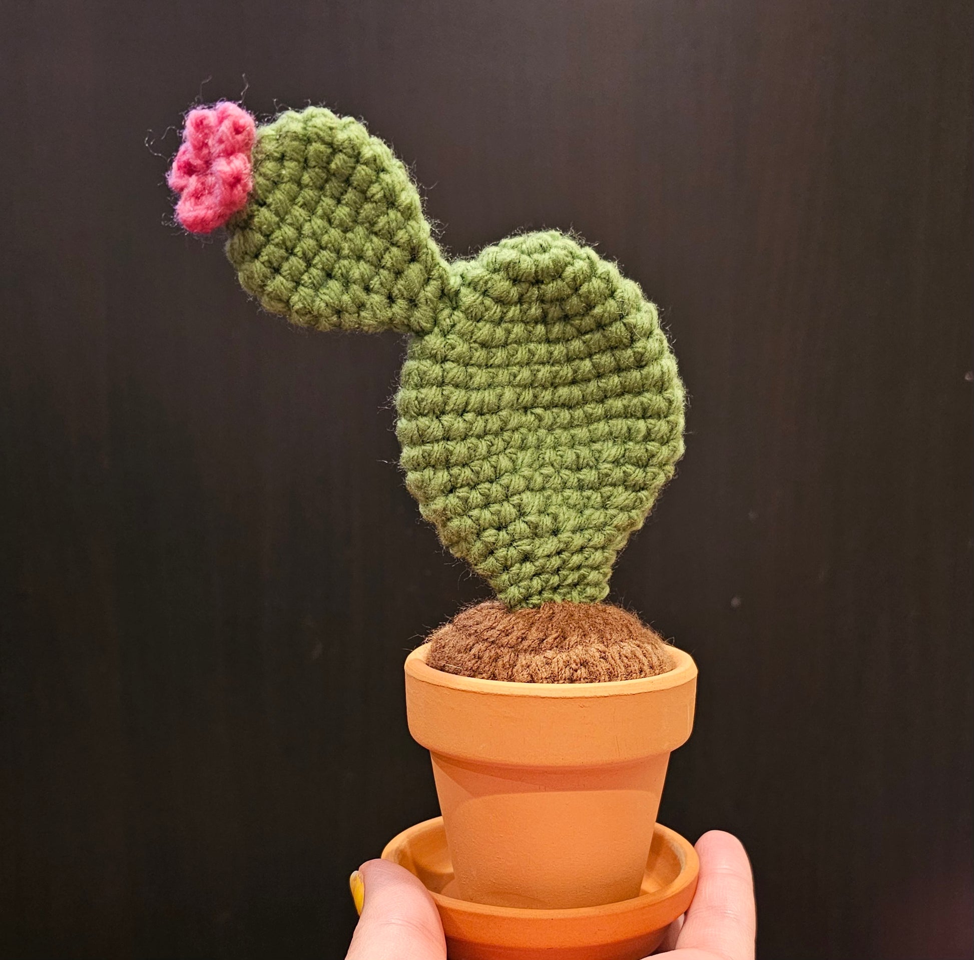 Crochet neutral green prickly pear cactus with a pink flower growing out of the top. The cactus is growing out of crochet dirt made out of brown yarn. The cactus sits in a small terracotta clay pot and saucer.