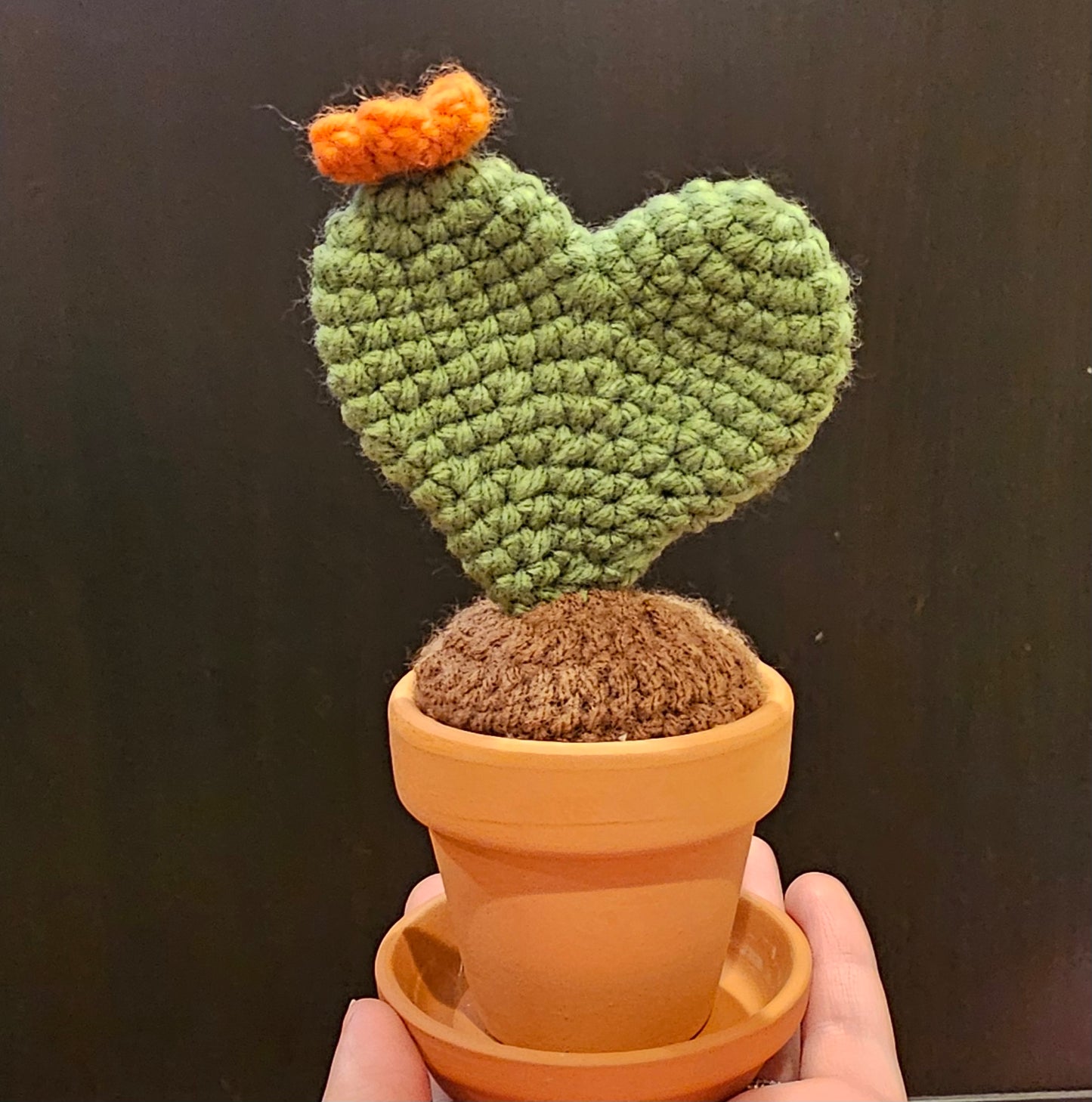 Crochet neutral green prickly pear cactus in the shape of a heart with an orange flower growing out of the top. The cactus is growing out of crochet dirt made out of brown yarn. The cactus sits in a small terracotta clay pot and saucer.