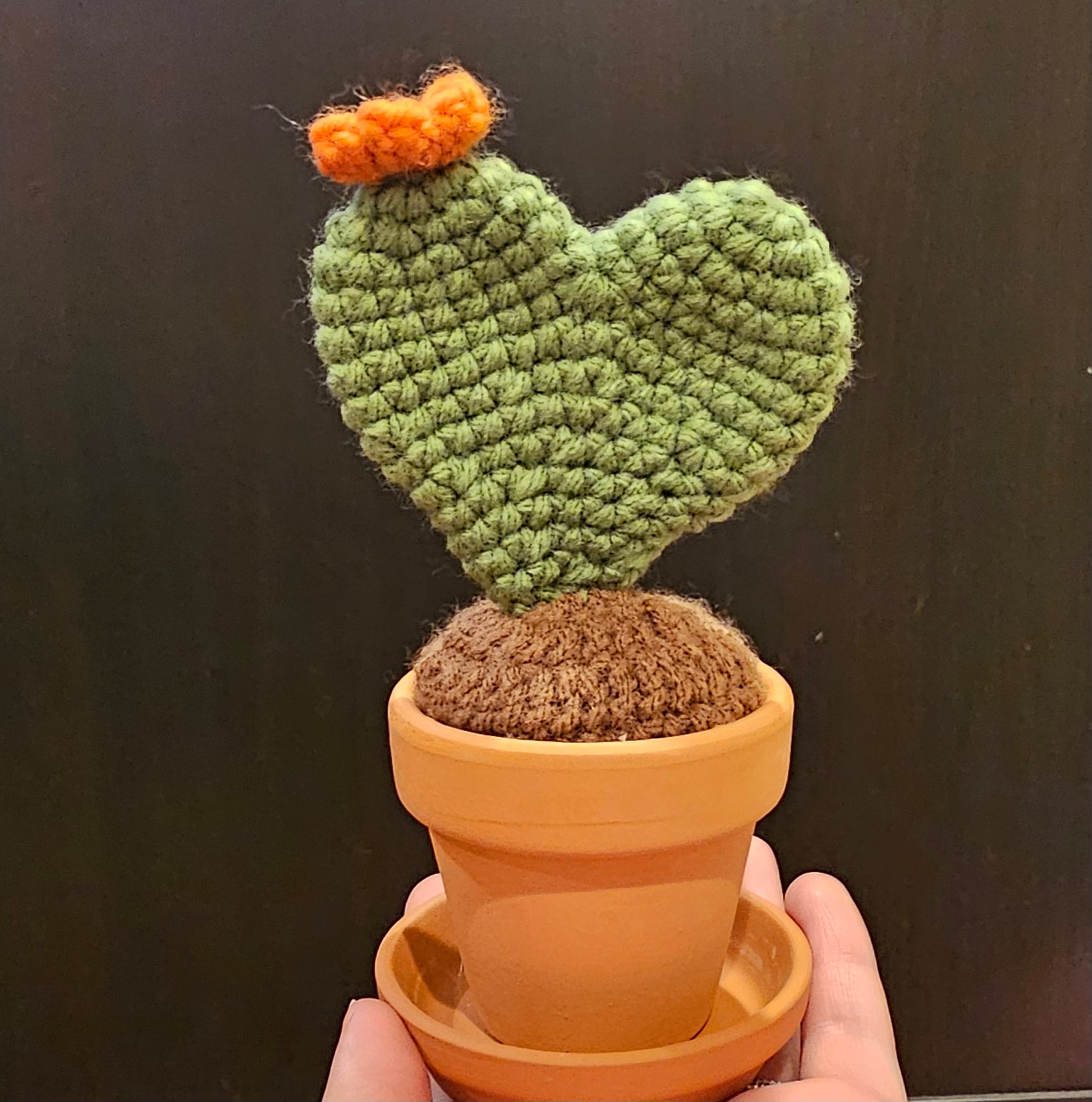 Crochet neutral green prickly pear cactus in the shape of a heart with an orange flower growing out of the top. The cactus is growing out of crochet dirt made out of brown yarn. The cactus sits in a small terracotta clay pot and saucer.