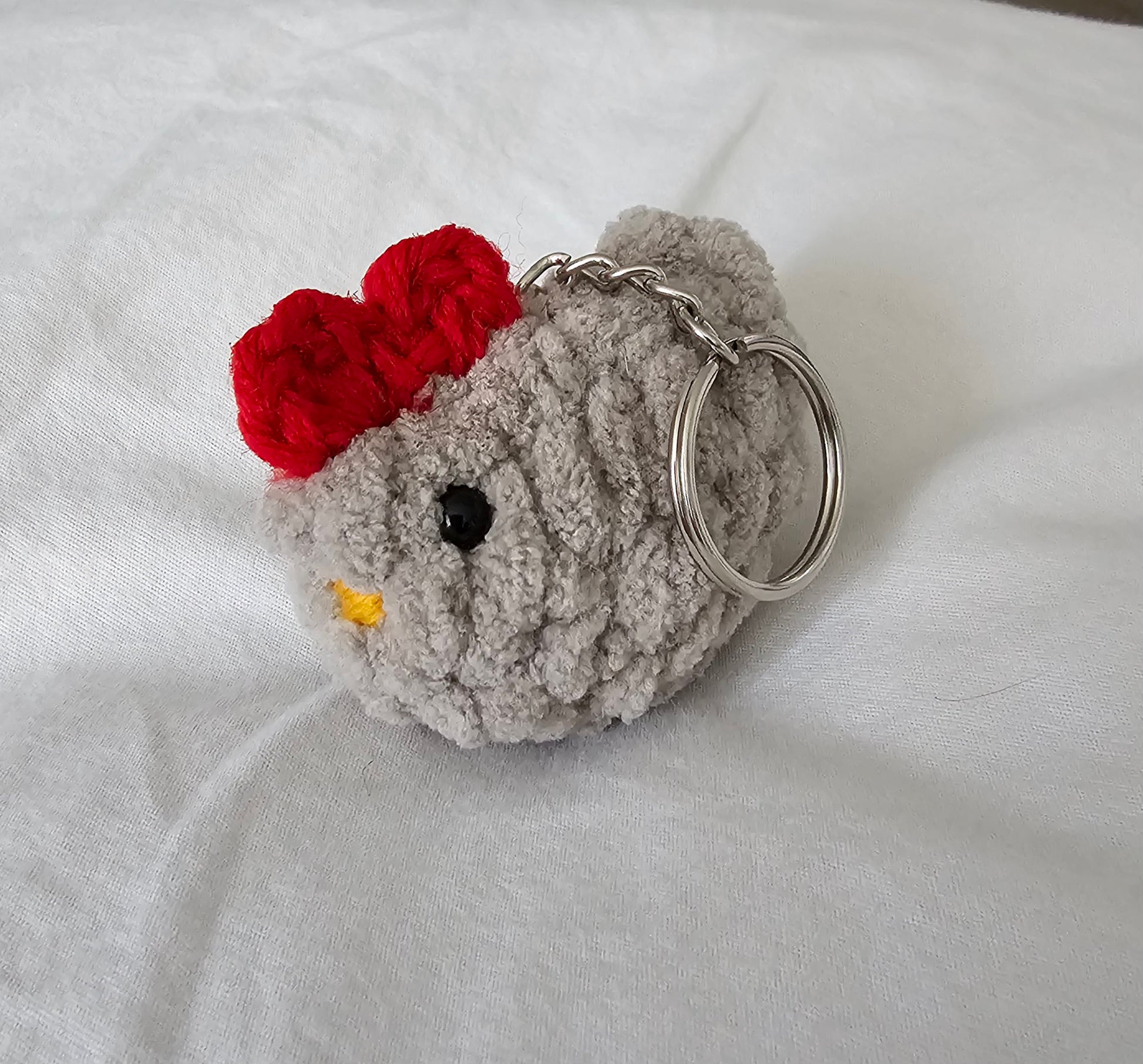 Small chicken keychain made from tan plush yarn. The keychain also has small black eyes, a bright yellow yarn beak and a red comb on the top of the head. The key ring is attached to the chicken by a small metal chain.