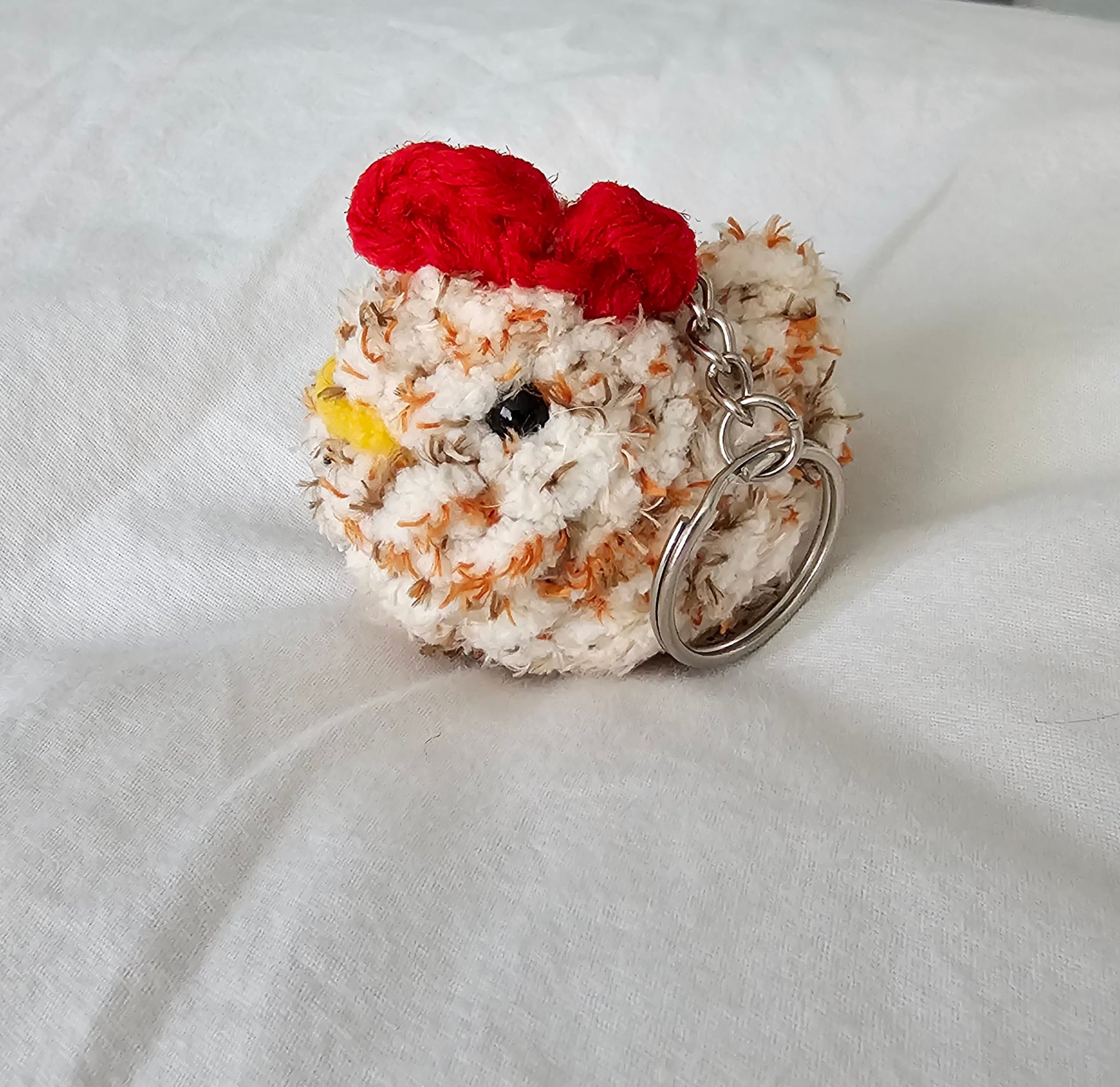 Small chicken keychain made from white and orange plush yarn. The keychain also has small black eyes, a bright yellow yarn beak and a red comb on the top of the head. The key ring is attached to the chicken by a small metal chain.