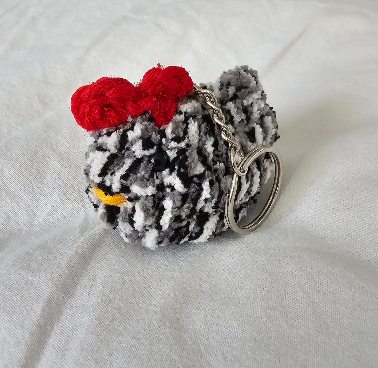 Small chicken keychain made from a yarn mix of white, black, and gray yarn. The keychain also has small black eyes, a bright yellow yarn beak and a red comb on the top of the head. The key ring is attached to the chicken by a small metal chain.