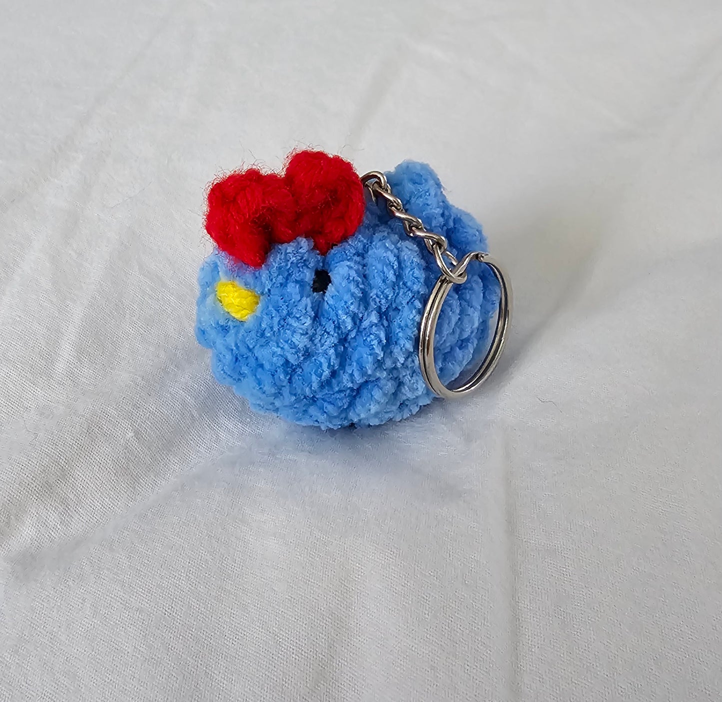 Small chicken keychain made from blue plush yarn. The keychain also has small black eyes, a bright yellow yarn beak and a red comb on the top of the head. The key ring is attached to the chicken by a small metal chain.