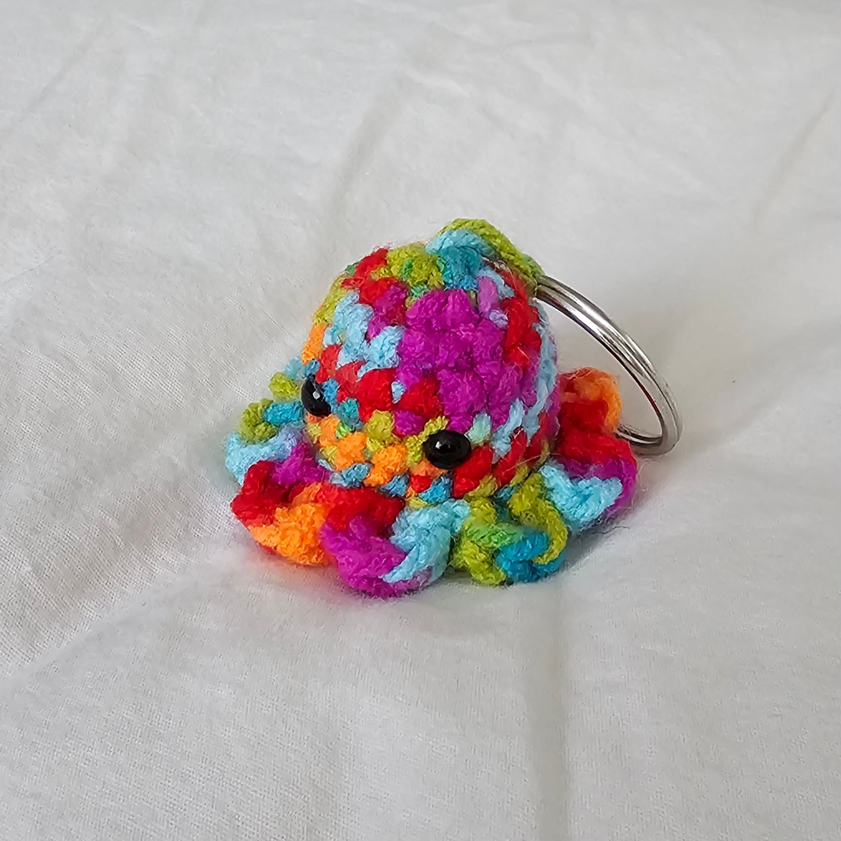 Small octopus keychain made from a multi color yarn which has a mix of bright rainbow colors (blue, green, red, orange, fuchsia) . The octopus also has small black eyes and the key ring is attached through the top of the head by the same yarn the octopus is made out of.