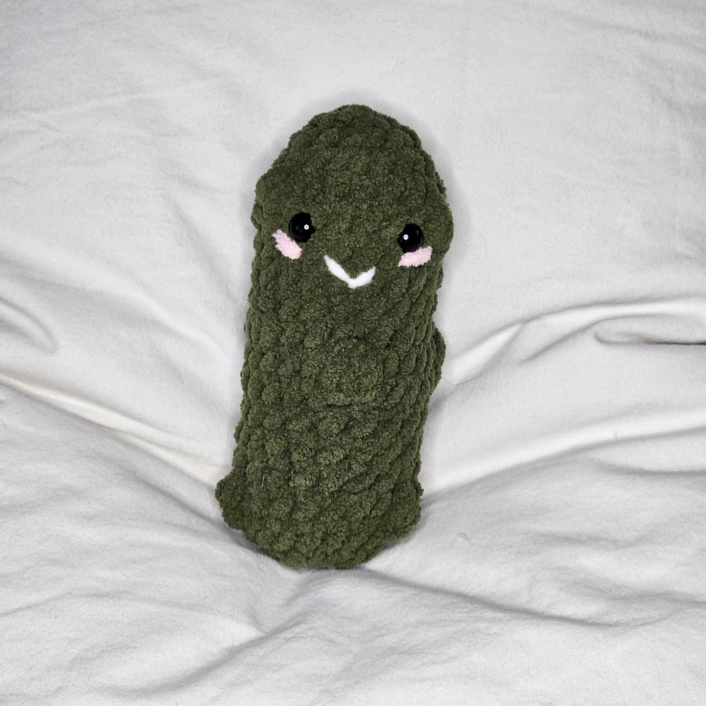 A small pickle made from olive green, plushie yarn. the pickle has small lumps throughout. it has little black eyes and pink yarn that runs just under the eyes as if it were blushing. It has a small smile made out of white yarn between the eyes.