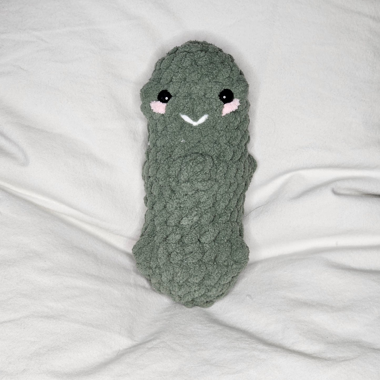 A small pickle made from pale green, plushie yarn. the pickle has small lumps throughout. it has little black eyes and pink yarn that runs just under the eyes as if it were blushing. It has a small smile made out of white yarn between the eyes.