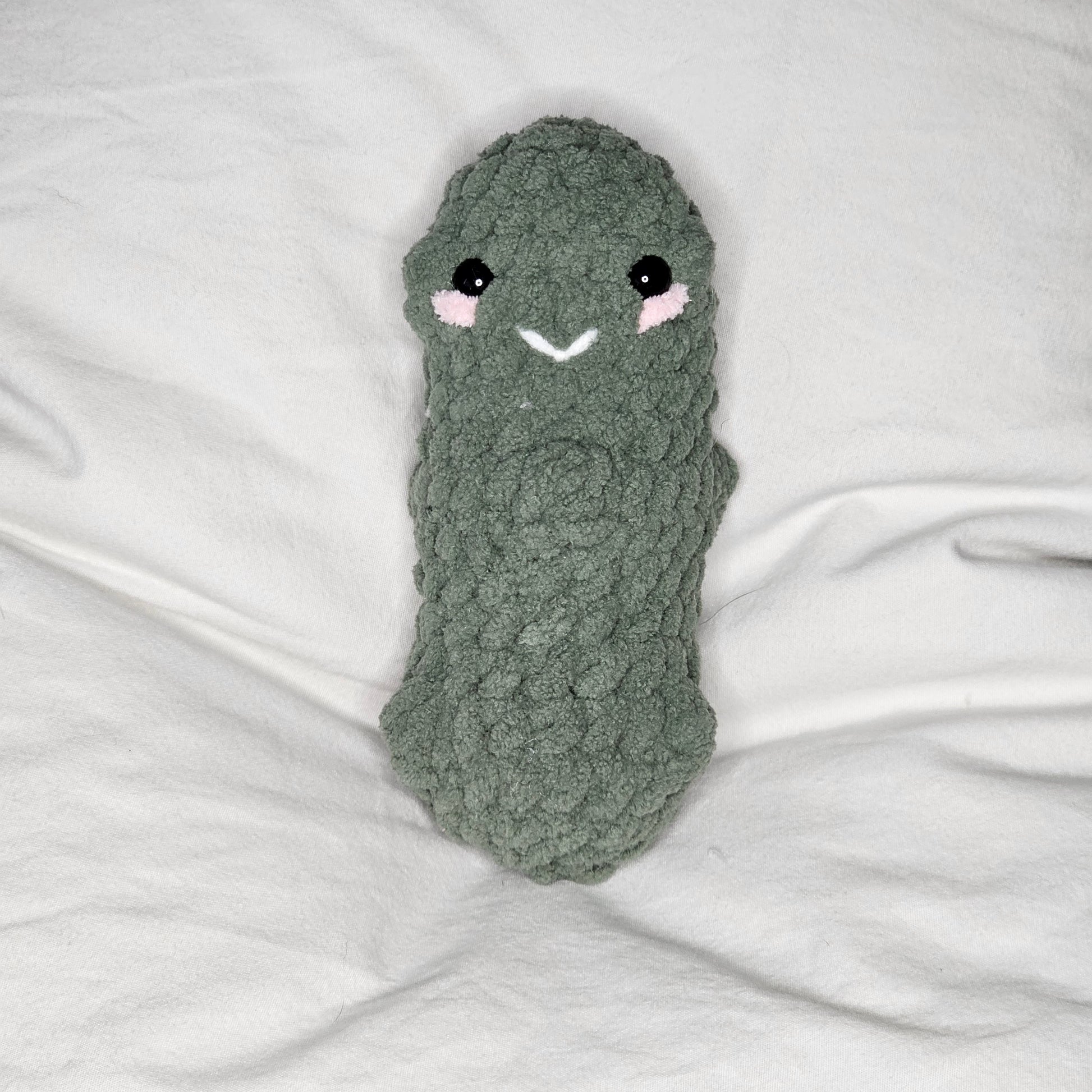 A small pickle made from pale green, plushie yarn. the pickle has small lumps throughout. it has little black eyes and pink yarn that runs just under the eyes as if it were blushing. It has a small smile made out of white yarn between the eyes.