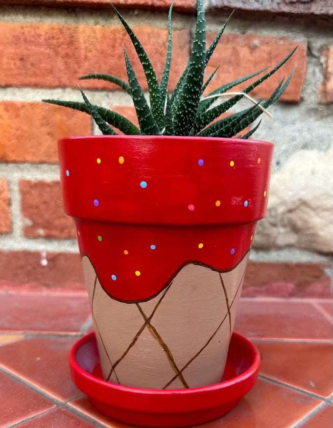 A tall decorative pot sitting on a saucer with a spiky plant popping out of the top. The pot is decorated to look like a red strawberry sorbet with multicolored sprinkles melting over the sides of the cone. The saucer is also red like the sorbet.