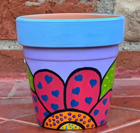 Decorative pot with pink flower petals that have blue hearts on them. There are green leaves behind the flower that have yellow sprinkles. The center of the flower is divided into two sections the very center is bright yellow with blue polka dots and the outer layer is orange with purple dots.The background is a lilac and the top ridge is painted pastel blue.