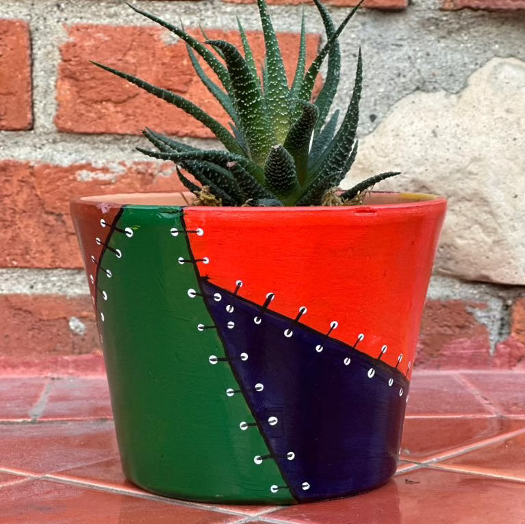 Multicolored decorative pot with patchwork design made to look like stitched on fabric. Each section is a different color, these colors are deeper tones like a rich orange, deep purple, warm brown, and forest green. There is a spiky plant sticking out of the top of the pot.