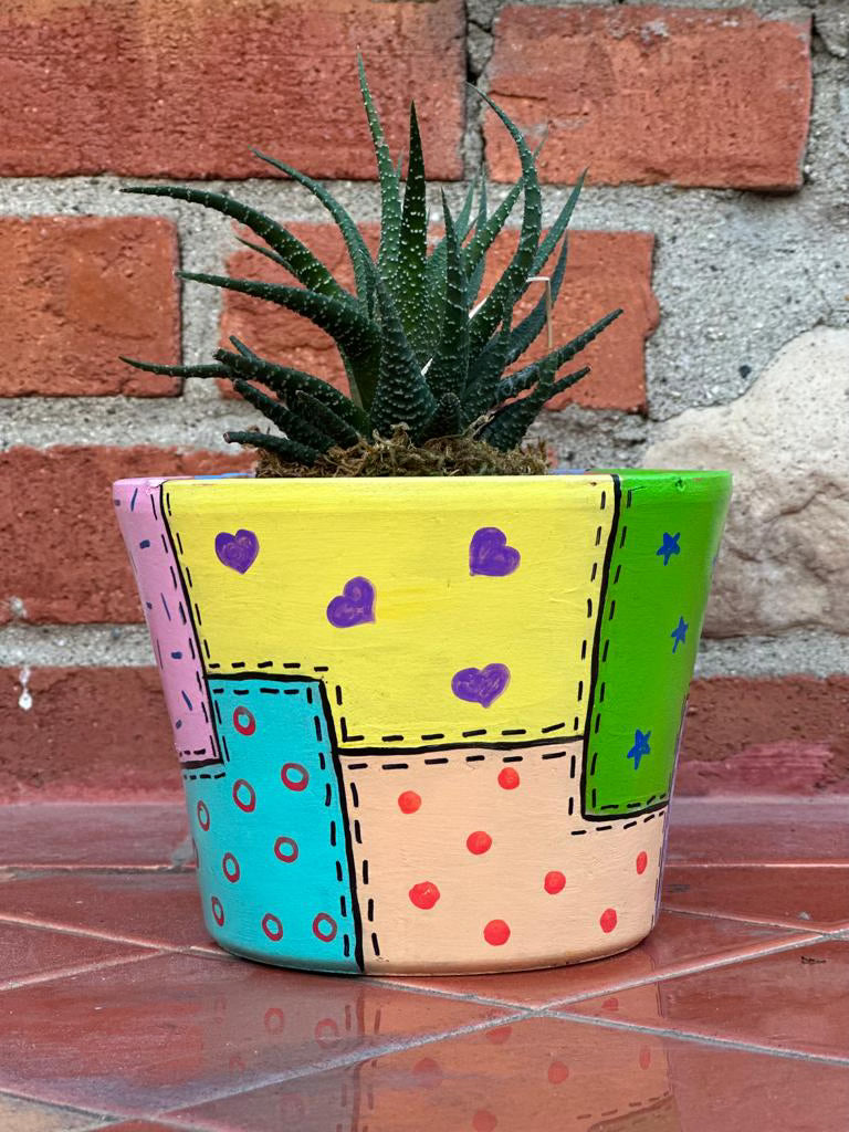 Multicolored decorative pot with patchwork design made to look like stitched on fabric. Each section is a different color and they are all pastels and lighter colors (yellow, blue, pink, peach, green). Each section is also decorated with smaller shapers inside like hearts, dots, sprinkles and stars in colors that offset the color of the sections they're in. There is a spiky plant sticking out of the top of the pot.
