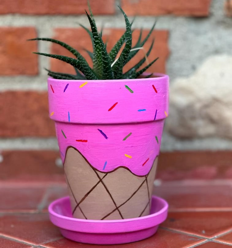 A tall decorative pot sitting on a saucer with a spiky plant popping out of the top. The pot is decorated to look like a pink strawberry ice cream with multicolored sprinkles melting over the sides of the cone. The saucer is also pink like the ice cream.