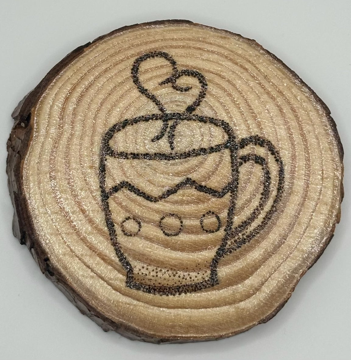 The disk on top has a design of a coffee cup. The cup has a zig zag line decorating it as well as three dots lining the bottoms half. There is steam leaving the cup in the shape of a heart burned onto the disk.
