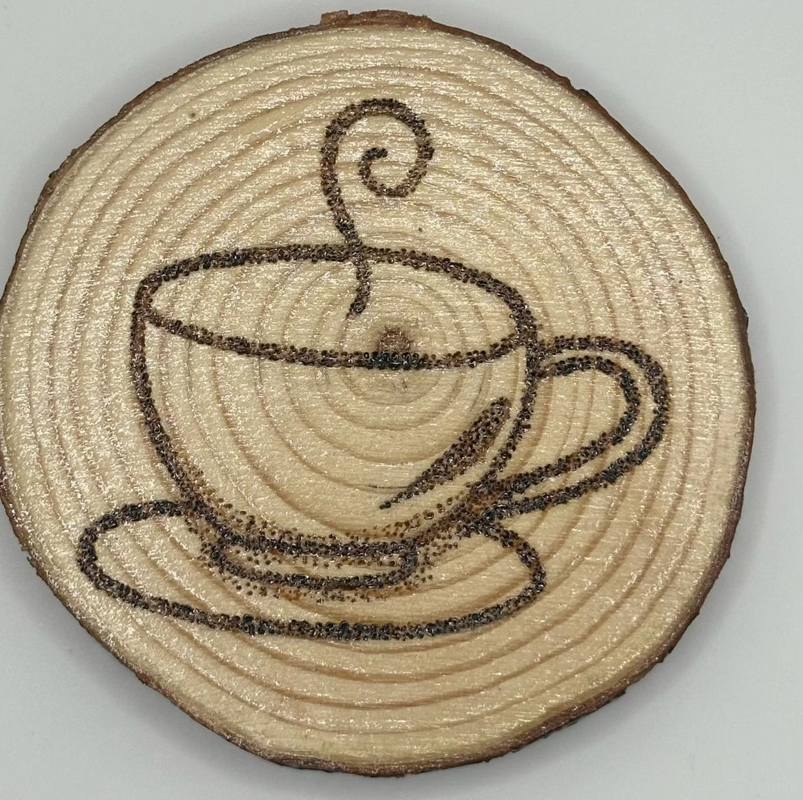 Wooden disk with a design burnt onto it. The design is a latte cup and saucer with a singular twirl of steam leaving the cup.
