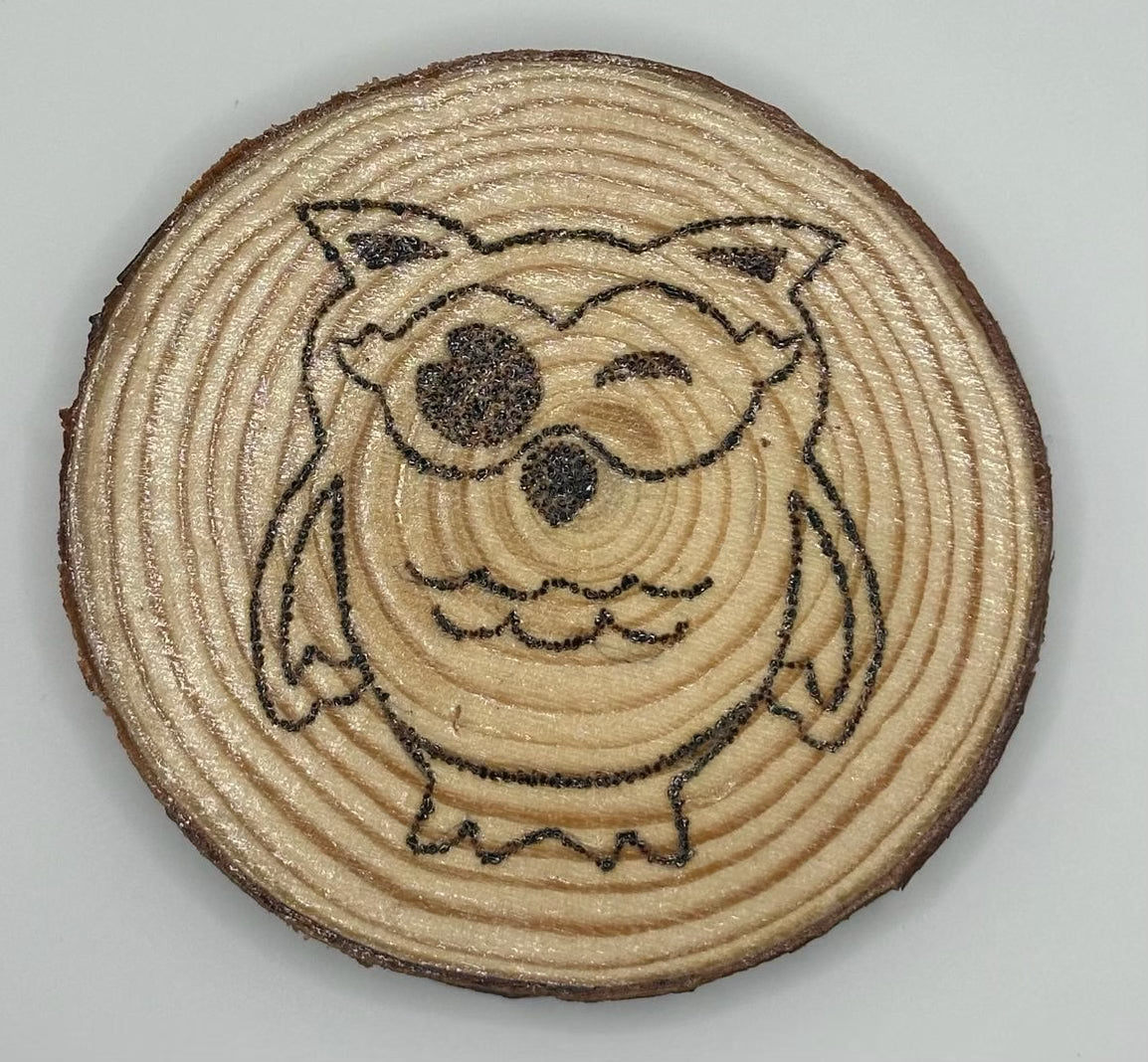 Wooden disk with a burnt on design. The design is a cute, big eyed owl that is blinking his right eye
