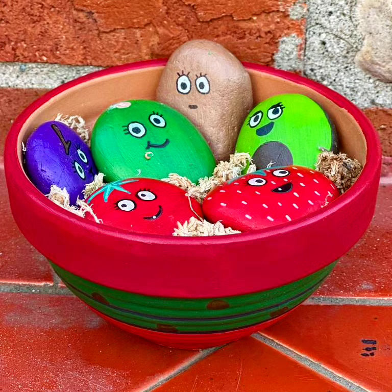 A bright red, bowl shaped, pot, with a green stripe running through the middle with little black dots scattered throughout. Inside the bowl, there are six rocks painted like different fruits and vegetables. There is a bright red strawberry and tomato, an avocado, a lime, a grape and a potato. All the rocks have different expressions between smiling excitement and surprise.