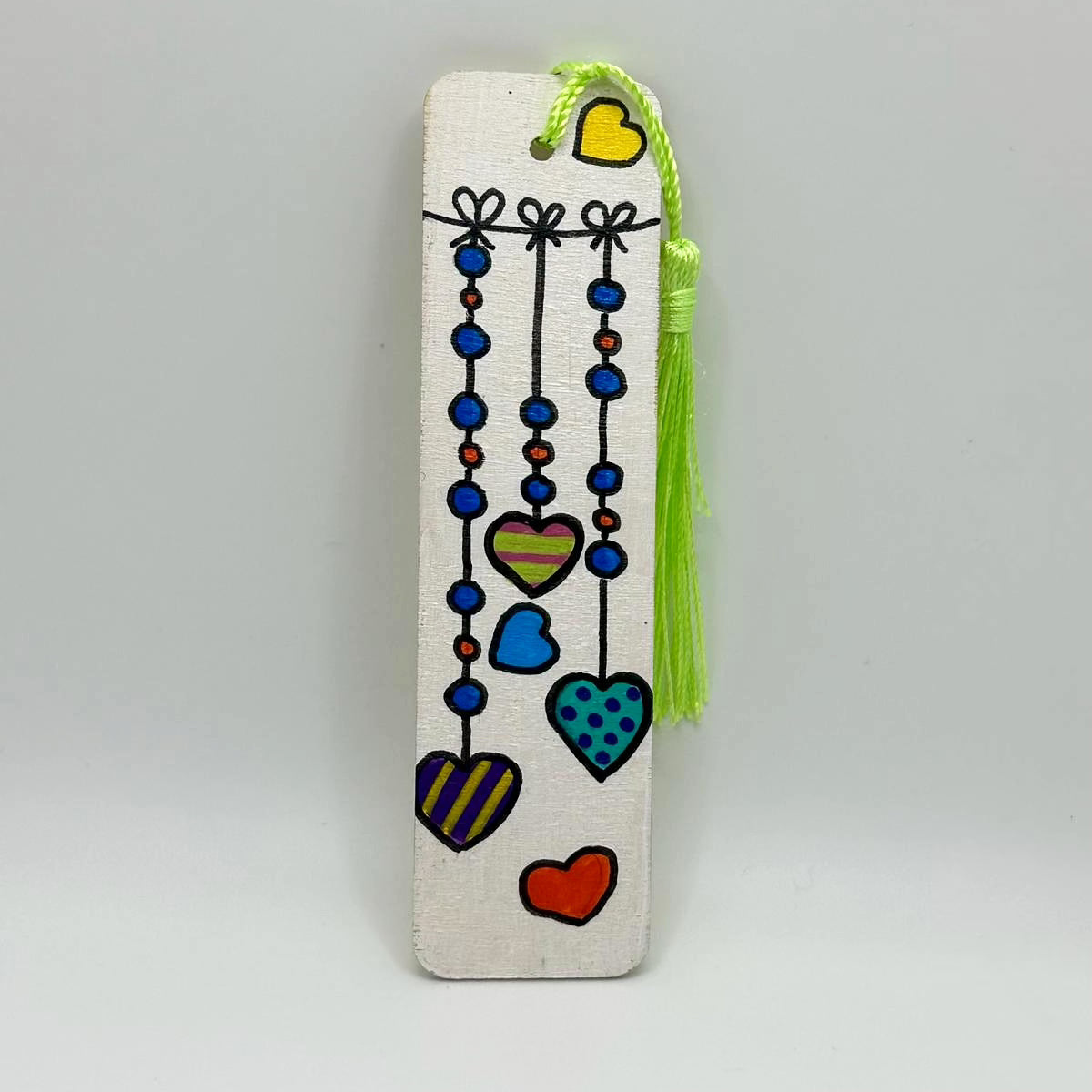 Small, white, wooden bookmark decorated with painted on hearts. The hearts are painted to look like they are hanging by strings with beads on them. The hearts are multicolored with varying patterns such as stripes and dots. The bookmark has a bright green tassel attached at the top through a small hole.