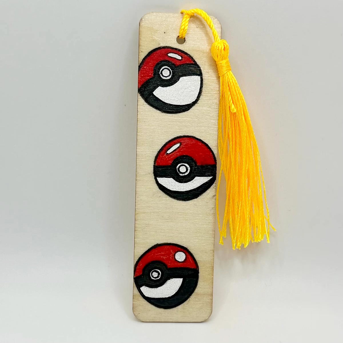 Small wooden bookmark decorated with painted on pokeballs that are red on top and white on bottom with a black stripe through the middle and a white button in the center. The bookmark has a bright yellow tassel attached at the top through a small hole.