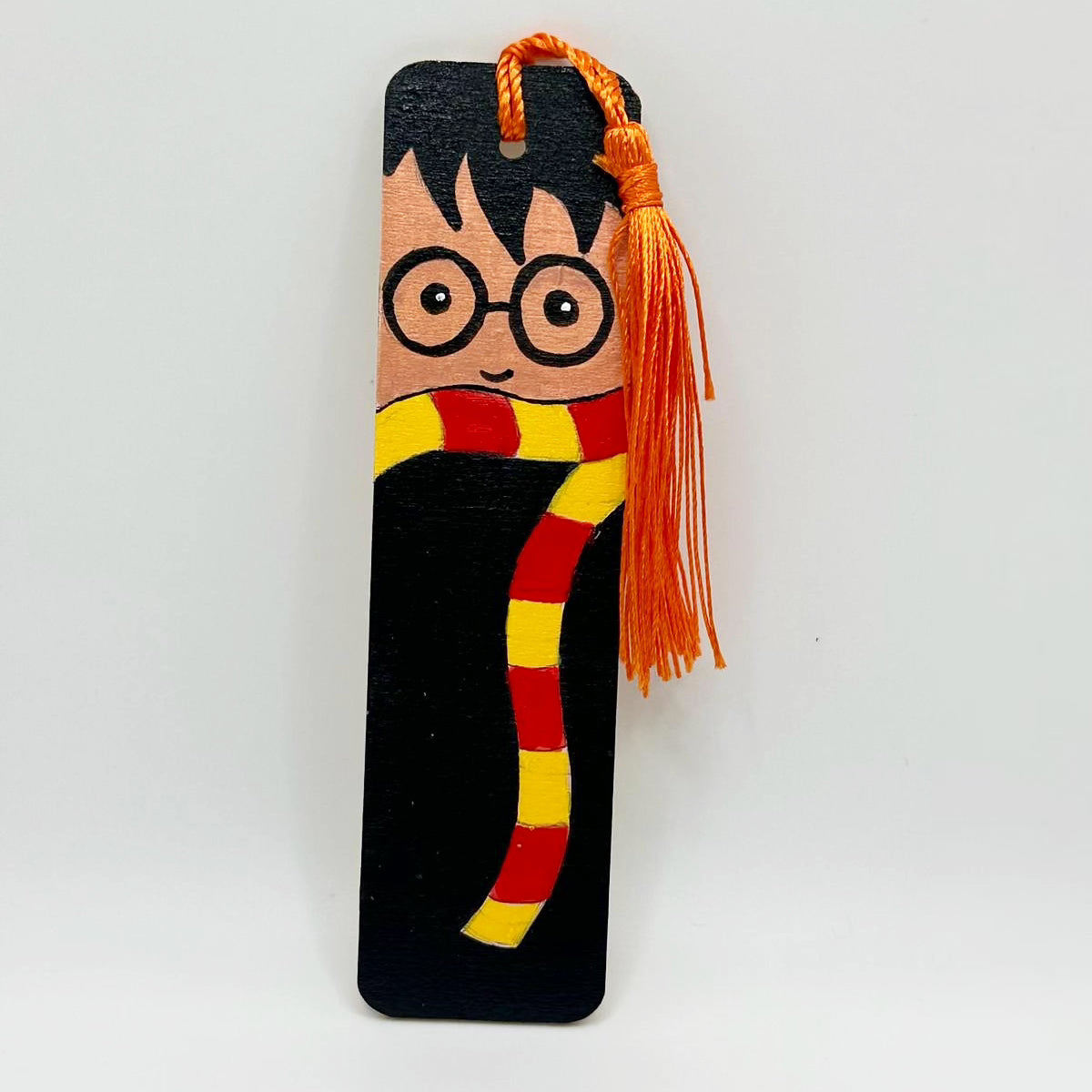 Small wooden bookmark decorated with a painted boy with black droopy hair and round eyeglasses. the boy wears a striped yellow and red scarf. The The bookmark has a bright orange tassel attached at the top through a small hole.