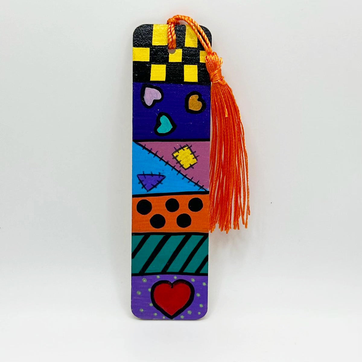 Small wooden bookmark painted with colorful designs such as checkers, stripes, polkadots, hearts and stitched on patchwork. There is a bright orange tassel attached to the bookmark through a small hole at the top of it.
