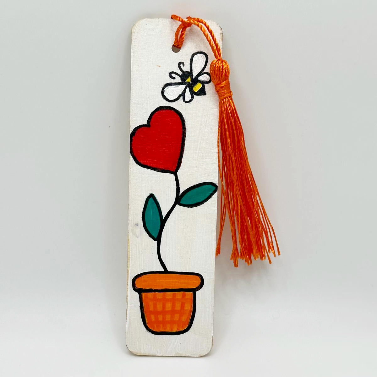 Small, white, wooden bookmark decorated with a red heart shaped flower growing out of an orange flower pot. There is a yellow and black bee with big white wings floating near the flower. The The bookmark has a bright orange tassel attached at the top through a small hole.