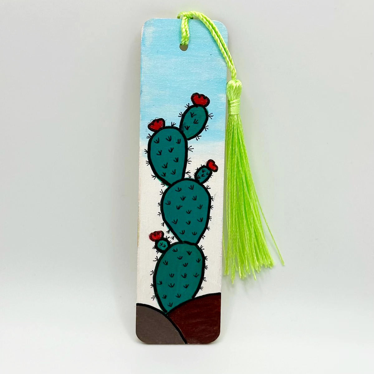 Small wooden bookmark with a blue sky background. There is a prickly pear cactus growing out of two small mounts of dirt. The cactus is four prickly pears tall with small red flowers growing out of it throughout. The cactus has many small thorns all over. There is a bright green tassel attached to the bookmark through a whole in the center.