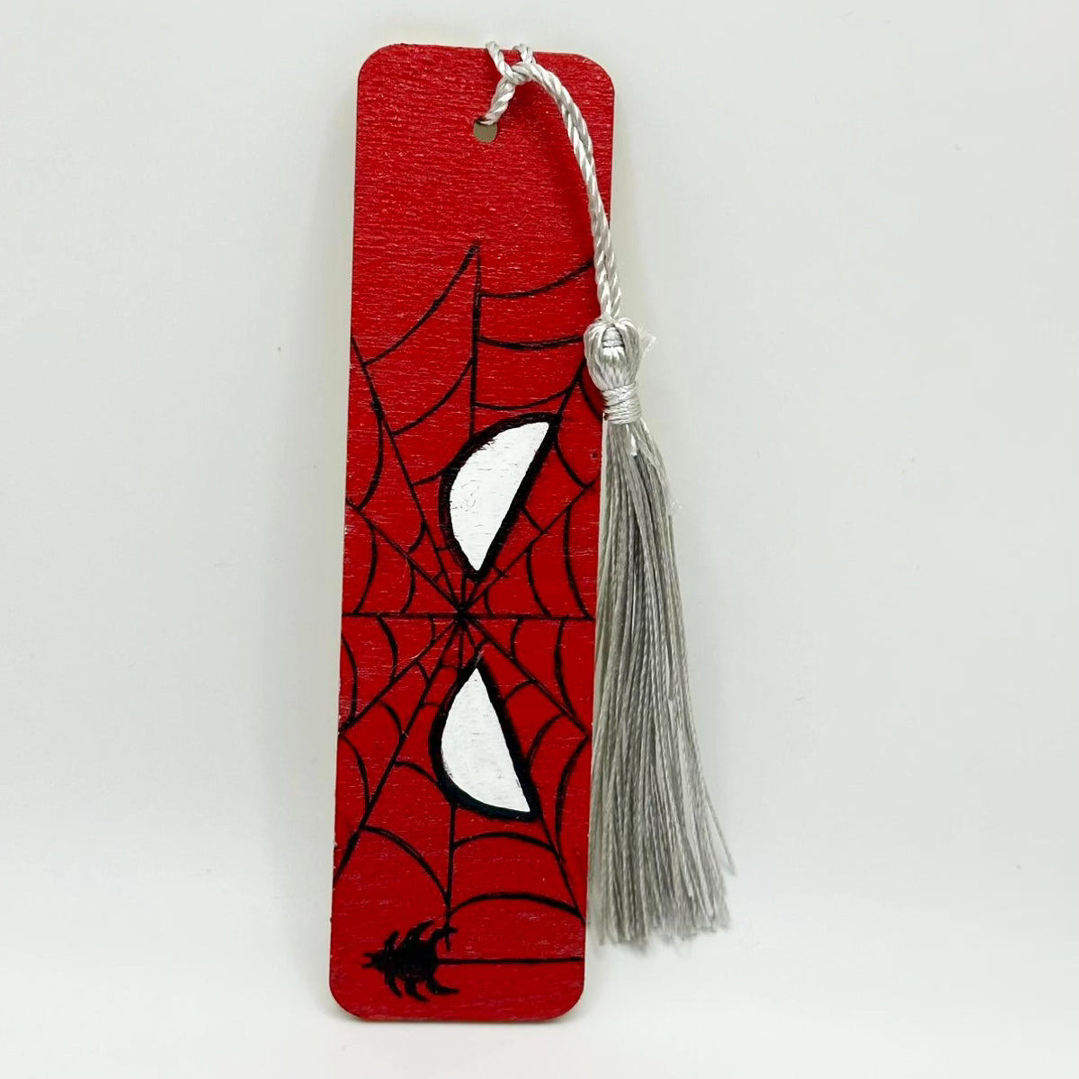Small, red, wooden bookmark with a drawn outline of the shape of white eyes over a spider web. There is a small black spider on the bottom left corner hanging by its spiderweb.The bookmark has a silver tassel attached at the top through a small hole.
