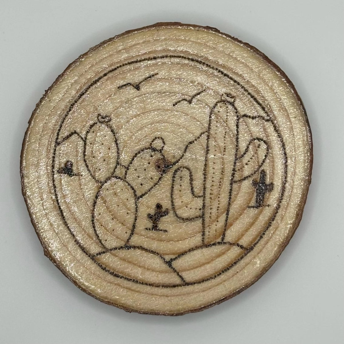 Wooden disk with a design burned onto it, the design is a landscape with some cacti up close and some far away. There are also some birds flying on the skyline. The landscape is encased in a circle.