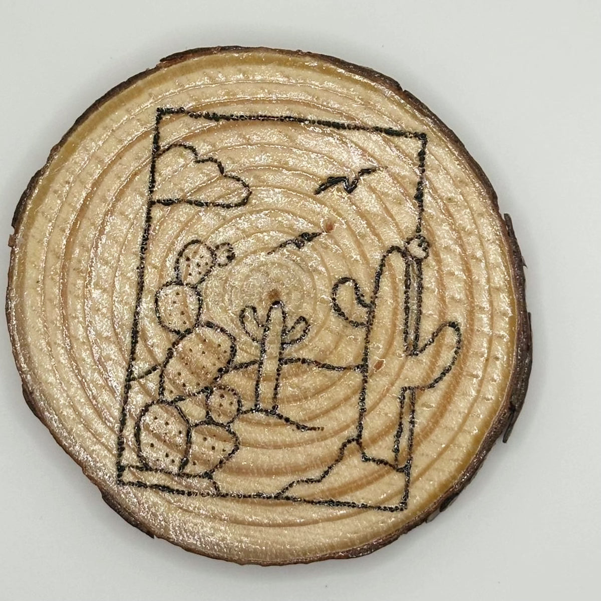 Wooden disk with a design burned onto it, the design is a landscape with some cacti up close and some far away. There is a cloud and some birds flying on the skyline. The landscape is encased in a circle.