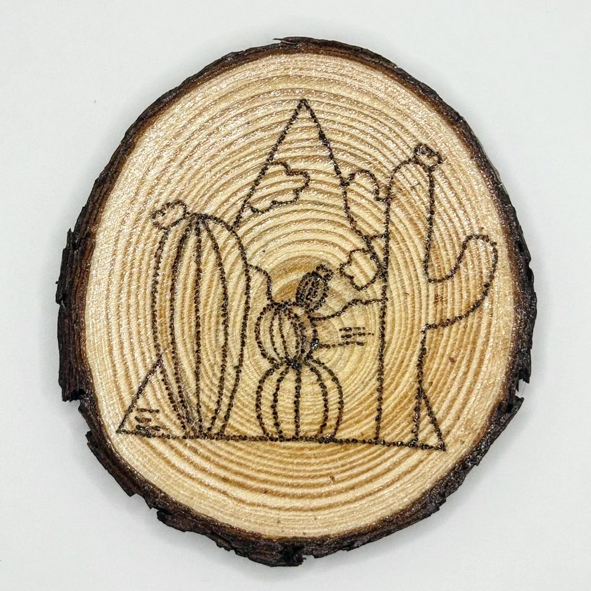 Wooden disk with a design burned onto it, the design is a landscape with some cacti up close and some far away. There are also some clouds on the skyline. The landscape is encased in a triangle but the cacti stand out of the lines of the triangle.