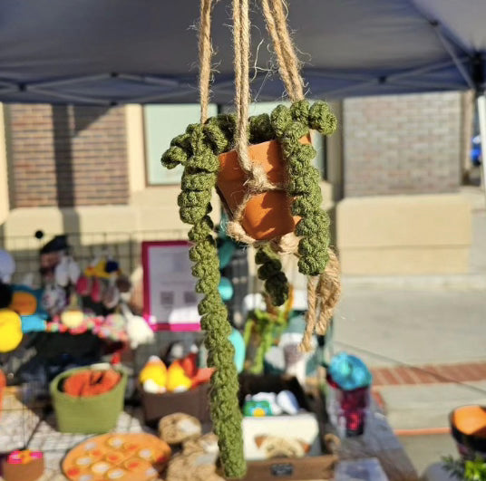 Spiraling plant crochet from olive green green yarn, hangs from a suspended terracotta pot. This pot sits in a jute rope macrame in front of a white background.